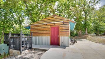 A small wooden cabin with a red door, surrounded by trees and a concrete patio.