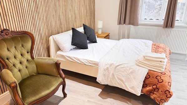 Habitación de hotel con una cama, cabecero de madera, sillón tapizado en verde, mesita de noche con lámpara y ventana con cortinas.