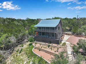 A two-story wooden house with a balcony, surrounded by a landscaped garden and dense forest. - Wimberley Hill Country Retreat | Sleeps 31, Firepits, Cabins & Scenic Views (Wimberley)