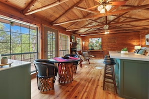 A rustic cabin interior with a wooden ceiling, large windows, a dining area with a table and chairs, a bar area, and a fireplace. - Wimberley Hill Country Retreat | Sleeps 31, Firepits, Cabins & Scenic Views (Wimberley)
