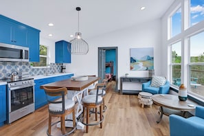 A modern kitchen with blue cabinets, a wooden dining table, and a large window. - Wimberley Hill Country Retreat | Sleeps 31, Firepits, Cabins & Scenic Views (Wimberley)