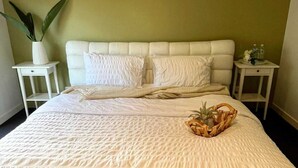 A neatly made bed with a white bedspread and pillows, two bedside tables with a vase and a bottle, and a potted plant on one side.