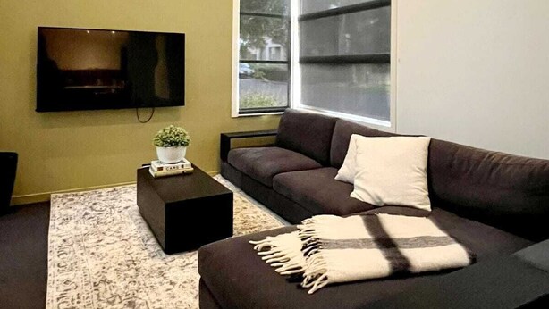 A modern living room with a large window, a flat-screen TV mounted on the wall, a dark sofa, a small coffee table, and a patterned rug.