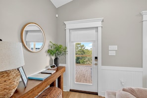 Une pièce avec un bureau en bois, un miroir, une plante en pot, une porte blanche donnant sur un balcon et une vue sur la verdure à l’extérieur.
