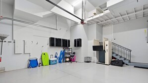 Un espace intérieur vaste et bien éclairé, avec des murs blancs, un escalier en métal et divers équipements, comme une planche de surf verte, une chaise pliante bleue et un meuble noir.