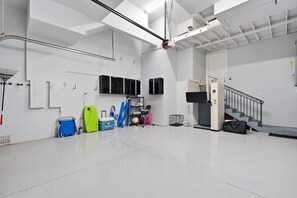 A spacious, well-lit indoor area with white walls, a metal staircase, and various equipment like a green surfboard, a blue folding chair, and a black cabinet. - Spacious Luxury Waterfront Retreat with Pool & Spa (Naples)