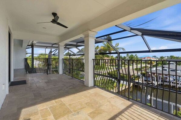 A covered balcony with a ceiling fan, a view of a street with palm trees, and a water body. - Spacious Luxury Waterfront Retreat with Pool & Spa (Naples)