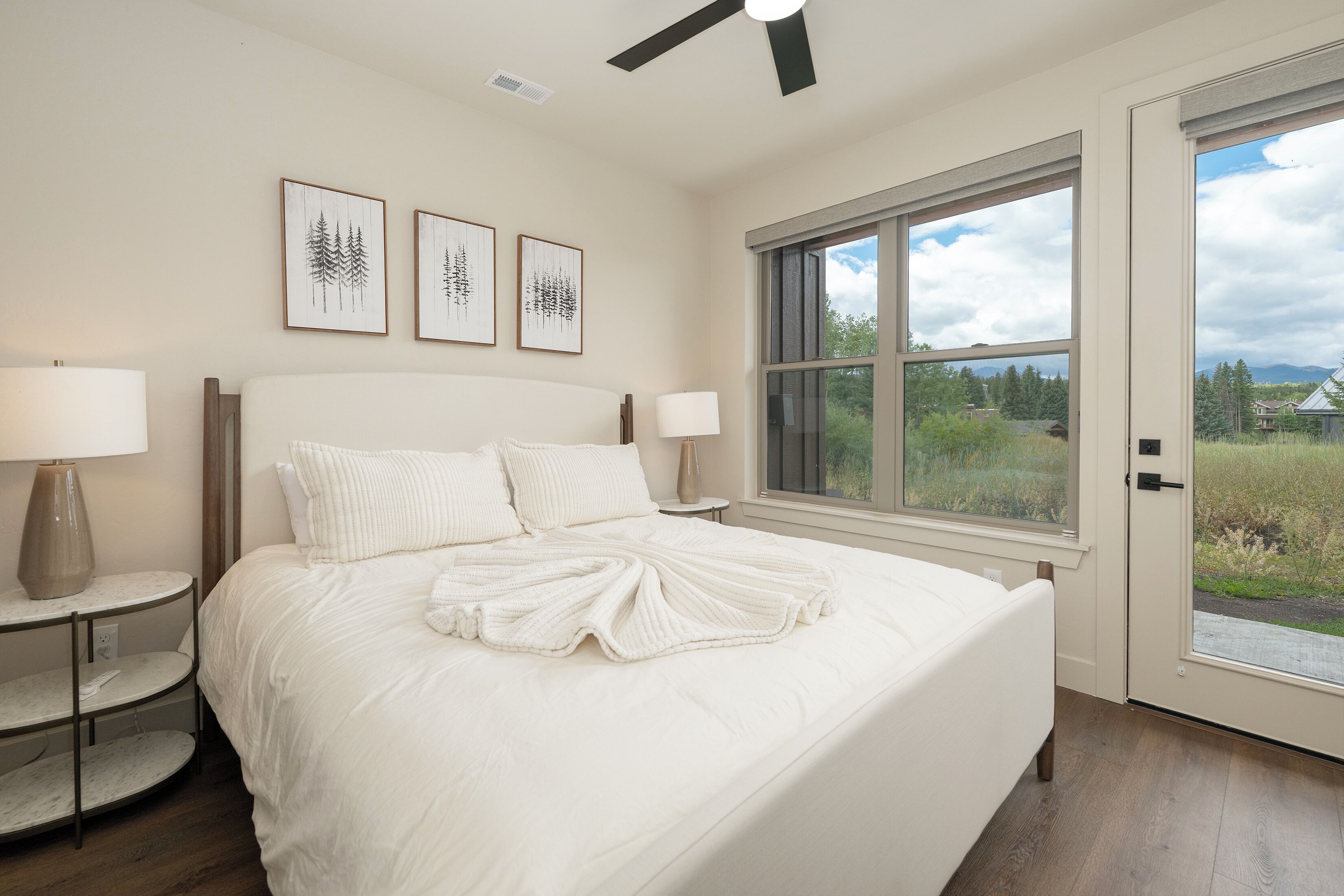 A bedroom with a large bed, two bedside tables with lamps, a ceiling fan, and a window with a view of greenery and mountains.
