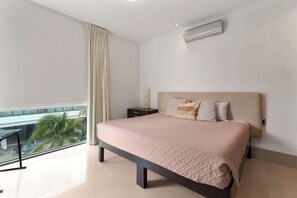 A bedroom with a bed, a nightstand, a window with a view of palm trees, and an air conditioning unit on the wall.