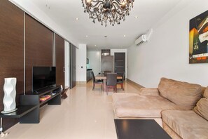 Un salon moderne avec un téléviseur à écran plat, un canapé beige, un coin repas avec une table et des chaises, et une cuisine en arrière-plan.