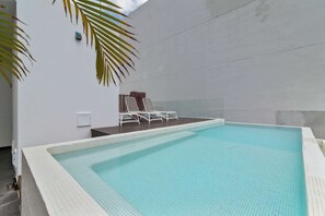 Ein Schwimmbecken mit gefliesten Rändern, einer weißen Wand und einer kleinen Sitzgelegenheit mit Stühlen und einer Bank.