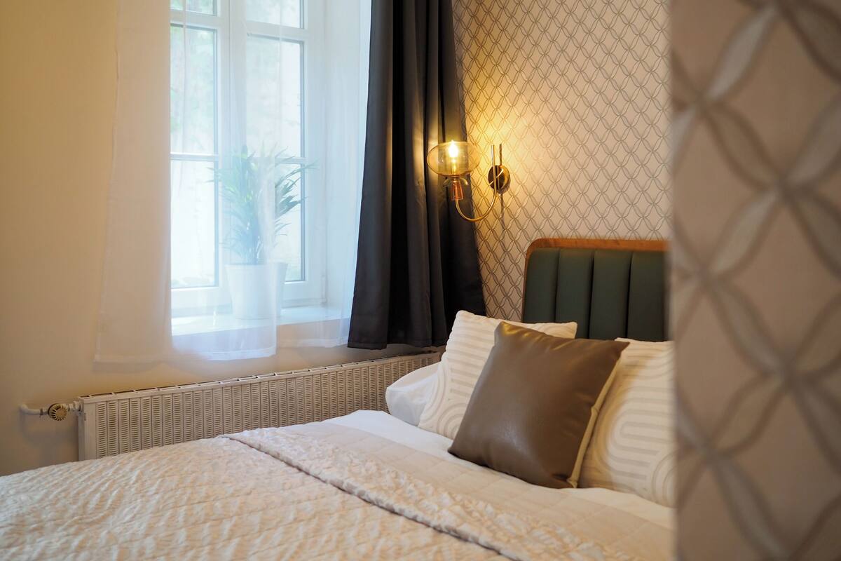 A neatly made bed with a quilt, a wall-mounted light fixture, and a window with curtains.