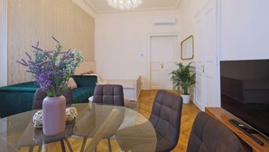 Una sala de estar moderna con una mesa de comedor de vidrio, sofás oscuros, una araña y un televisor de pantalla plana.