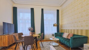 Una sala de estar moderna con un sofá verde, una mesa de comedor con sillas, un televisor en un mueble de madera y ventanas amplias con cortinas.