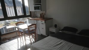A bedroom with a bed, a desk with a chair, a small bedside table, and a window with a view of buildings.