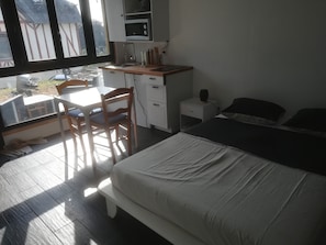 Un dormitorio con una cama, un escritorio con una silla, una mesita de noche pequeña y una ventana con vistas a edificios.