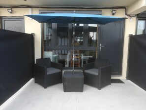 A patio area with two wicker chairs and a table, a blue umbrella, and a glass door leading to an interior space.