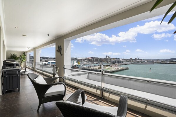 A balcony with wicker chairs, a small table, and a grill. It overlooks a marina with boats and buildings.