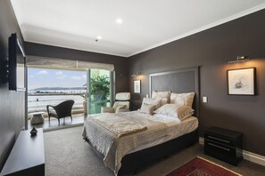 A modern bedroom with a large bed, a desk, and a chair. There is a balcony with a view of the sea.