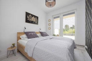 A bedroom with a bed, a small side table, a potted plant, a window with a view, and a wall-mounted artwork.