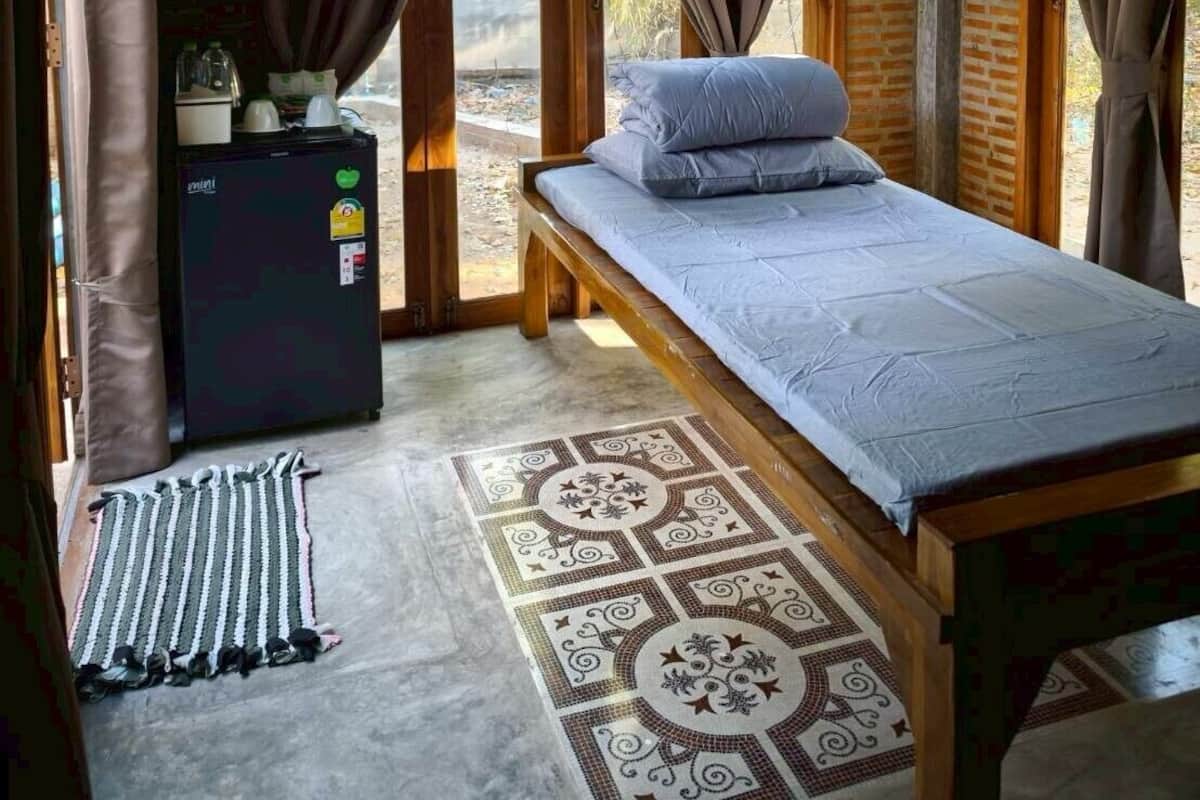 A single bed with a grey mattress and pillows, a small black refrigerator, a patterned rug, and a view of the outdoors through a wooden door.