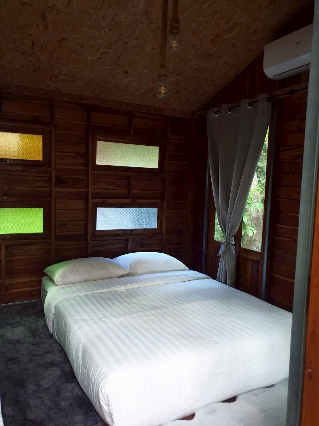 Una habitación de cabaña de madera con una cama, dos ventanas con persianas y un equipo de aire acondicionado.