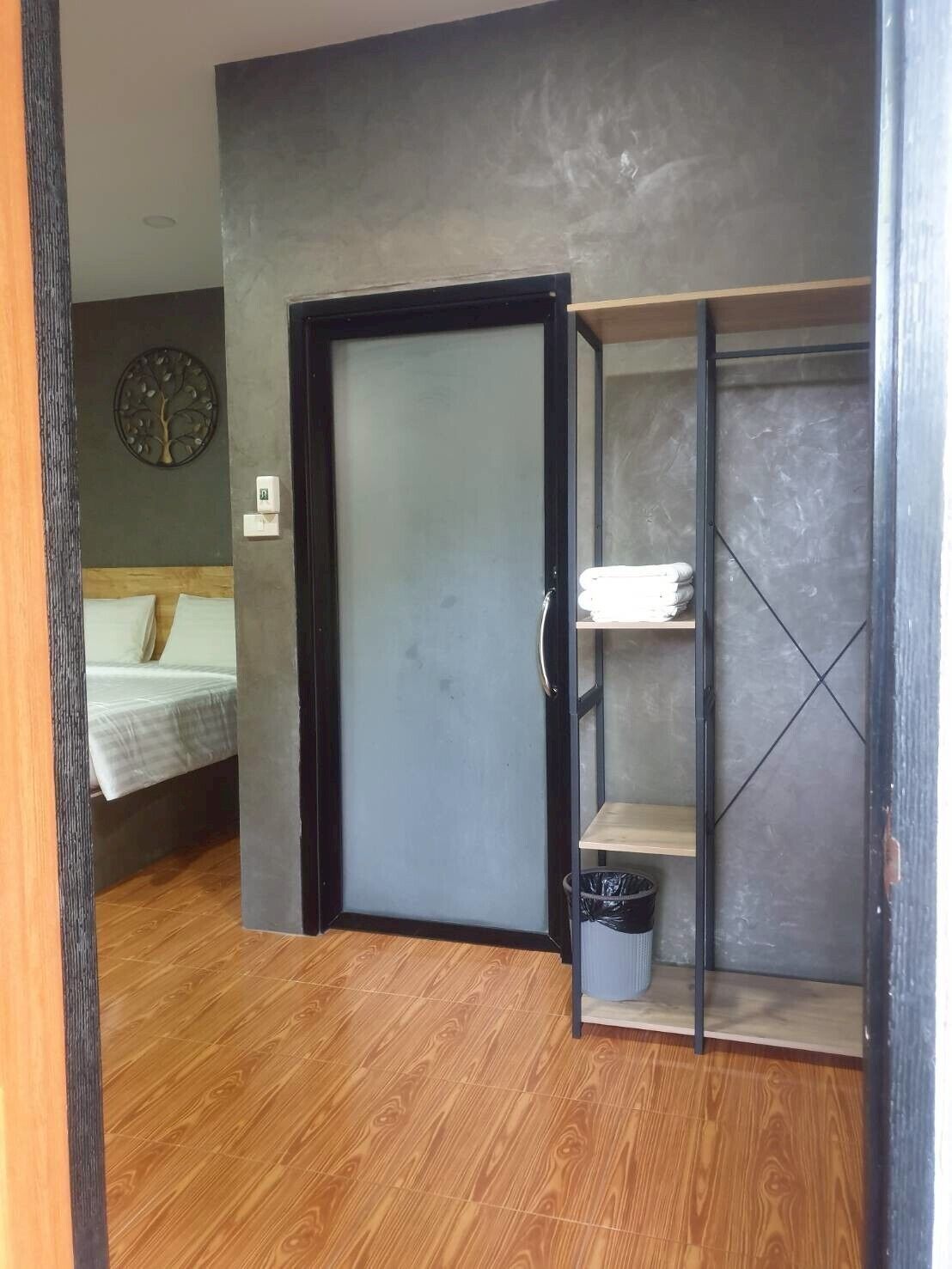 Una habitación con piso de madera, una cama con cubrecama blanco, una puerta gris y un bote de basura negro.
