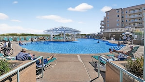 Zona de piscina con sillones de descanso, un espacio de estar cubierto y un edificio con balcones.
