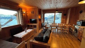 A cozy cabin interior with a wooden dining table, a sofa, a TV, and a view of snowy mountains through large windows.