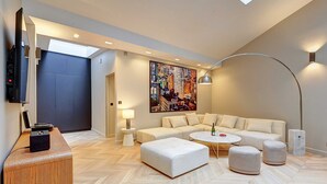 A modern living room with a sectional sofa, ottomans, a coffee table, a TV, and a painting on the wall. - Elegant Apartment With AC - 3br/10p- Le Marais (Paris)