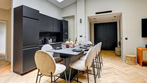 A modern kitchen with a large island, black cabinets, and a dining area with a long table and chairs. - Elegant Apartment With AC - 3br/10p- Le Marais (Paris)