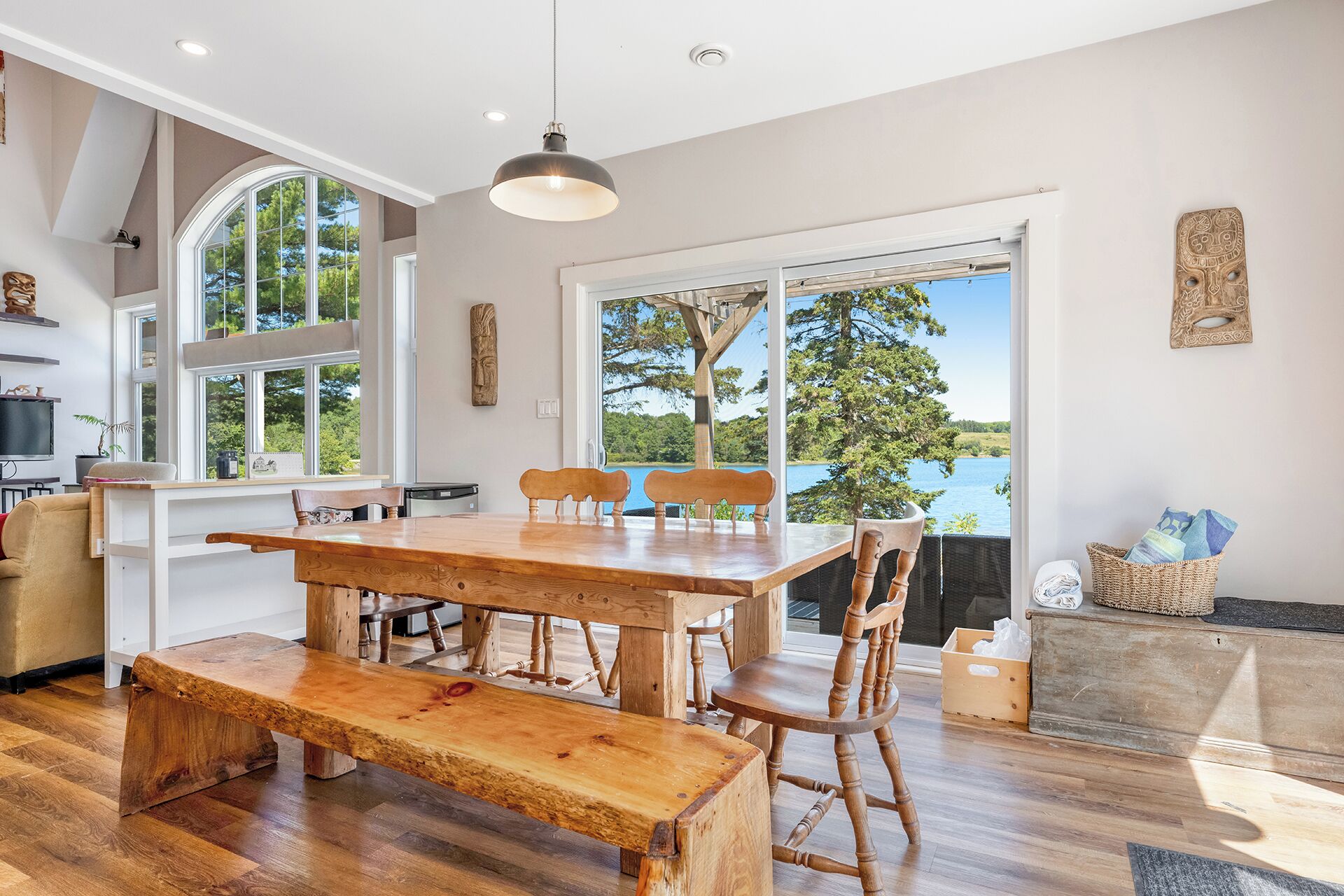 A spacious dining area with a wooden table and benches, large windows offering a view of a lake, and a cozy seating area with a wicker basket.