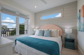 Ein Schlafzimmer mit einem großen Bett, zwei Fenstern, einem Balkon und einem Nachttisch mit einer Lampe.