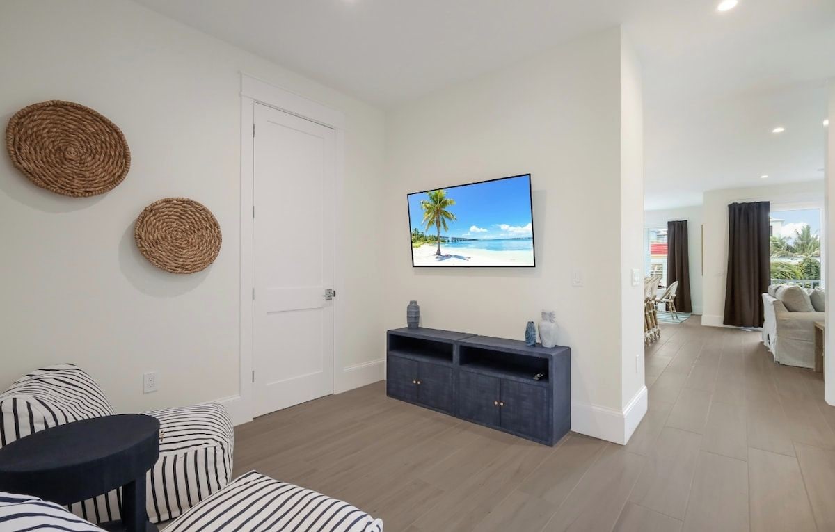 A modern living room with a sofa, a coffee table, a TV mounted on the wall, and a view of the beach through the window.