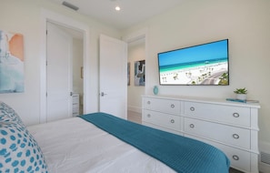 Ein Schlafzimmer mit einem Bett, einer Kommode, einem Fernseher, der eine Strandansicht zeigt, und einer Tür zu einem anderen Raum.