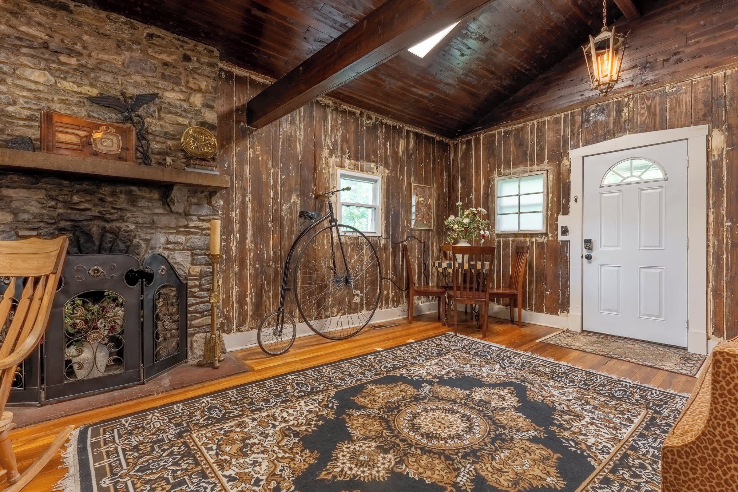 A rustic interior with a stone wall, wooden beams, a bicycle, a door, and a patterned rug.