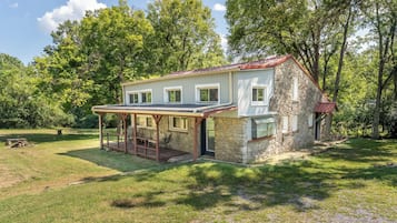 A stone house with a covered porch and a red roof, surrounded by a grassy yard and trees.
