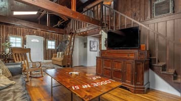 A rustic interior with a wooden staircase, a television on a wooden cabinet, and a dining table with a 'DOPE IN' sign.