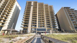A multi-story residential building with balconies, a central entrance, and a wooden walkway leading to a beach area.