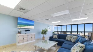 A spacious room with a large window, a blue sofa, a white cabinet, and a flat-screen TV mounted on the wall.
