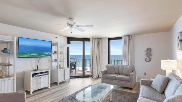 A living room with a television, a sofa, a glass coffee table, a ceiling fan, and a view of the ocean through a sliding door.