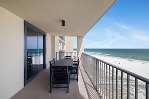 A balcony with a view of the beach and ocean, a table and chairs, and a sliding glass door.