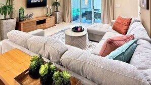Een moderne woonkamer met een grijze bank, een houten salontafel met planten, een televisie aan de muur en een groot raam met gordijnen.