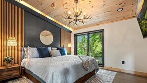 A bedroom with a large bed, a chandelier, a mirror, and a view of greenery outside.