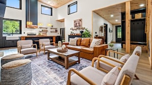 A spacious living room with a large sofa, armchairs, and ottomans, a coffee table, and a fireplace. There is a kitchen area with a dining table and chairs, and a large window providing a view of the outdoors.