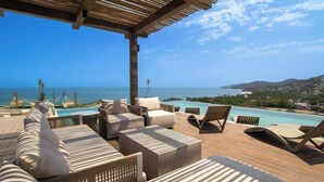 Terraza de madera con sillones y sofás con vistas a una piscina y al océano.