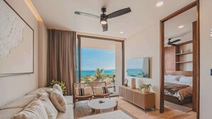 A modern hotel room with a sofa, a coffee table, a dining area, a bed, and a view of the ocean.