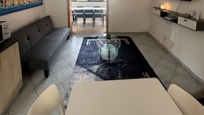 A modern living room with a sofa, dining table, and TV. A ceiling fan is visible, and there's a painting on the wall.