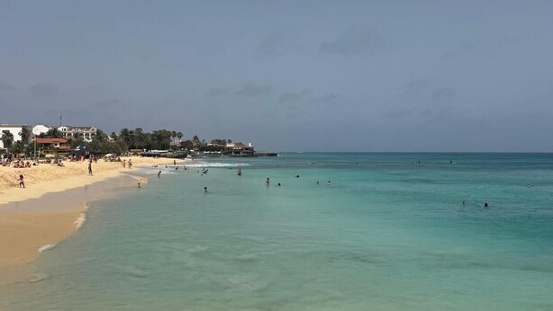 A beach with clear turquoise water, a sandy shore, and a few buildings in the background.