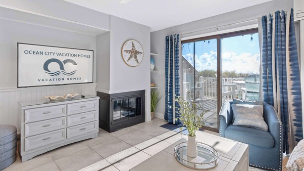 A modern living room with a fireplace, a large window with curtains, a sofa, and a wall-mounted sign for Ocean City Vacation Homes. - Seal Beach 2 (Ocean City)